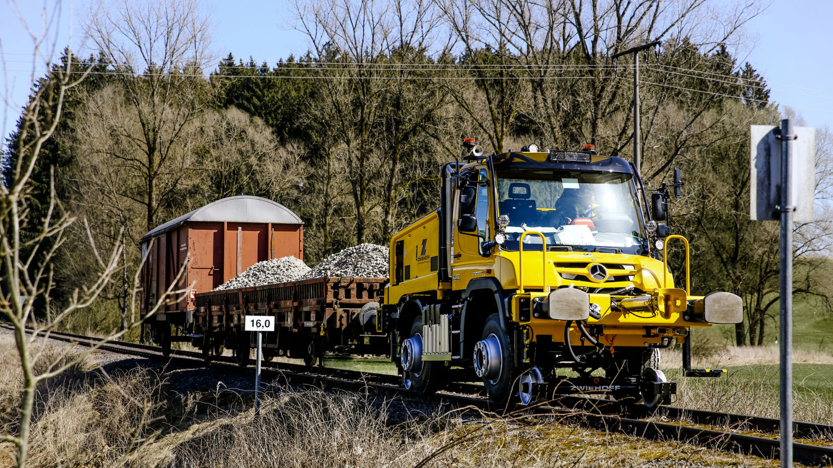 Kollane pealisehitusega Unimog, manööverdab kaubavaguneid rööbastel tööstusalal, taustal virnastatud betoonlävepakud ja kraanad.