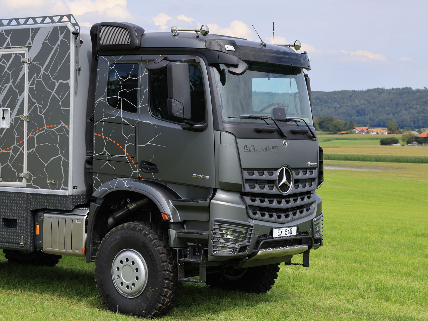 Das Bild zeigt einen grauen Arocs mit einem Expeditionsaufbau, der auf einer grünen Wiese steht. Das Fahrzeug hat ein markantes Design mit einem Muster, das an Risse oder Kartenlinien erinnert. Im Hintergrund sind Felder, Häuser und bewaldete Hügel zu sehen.