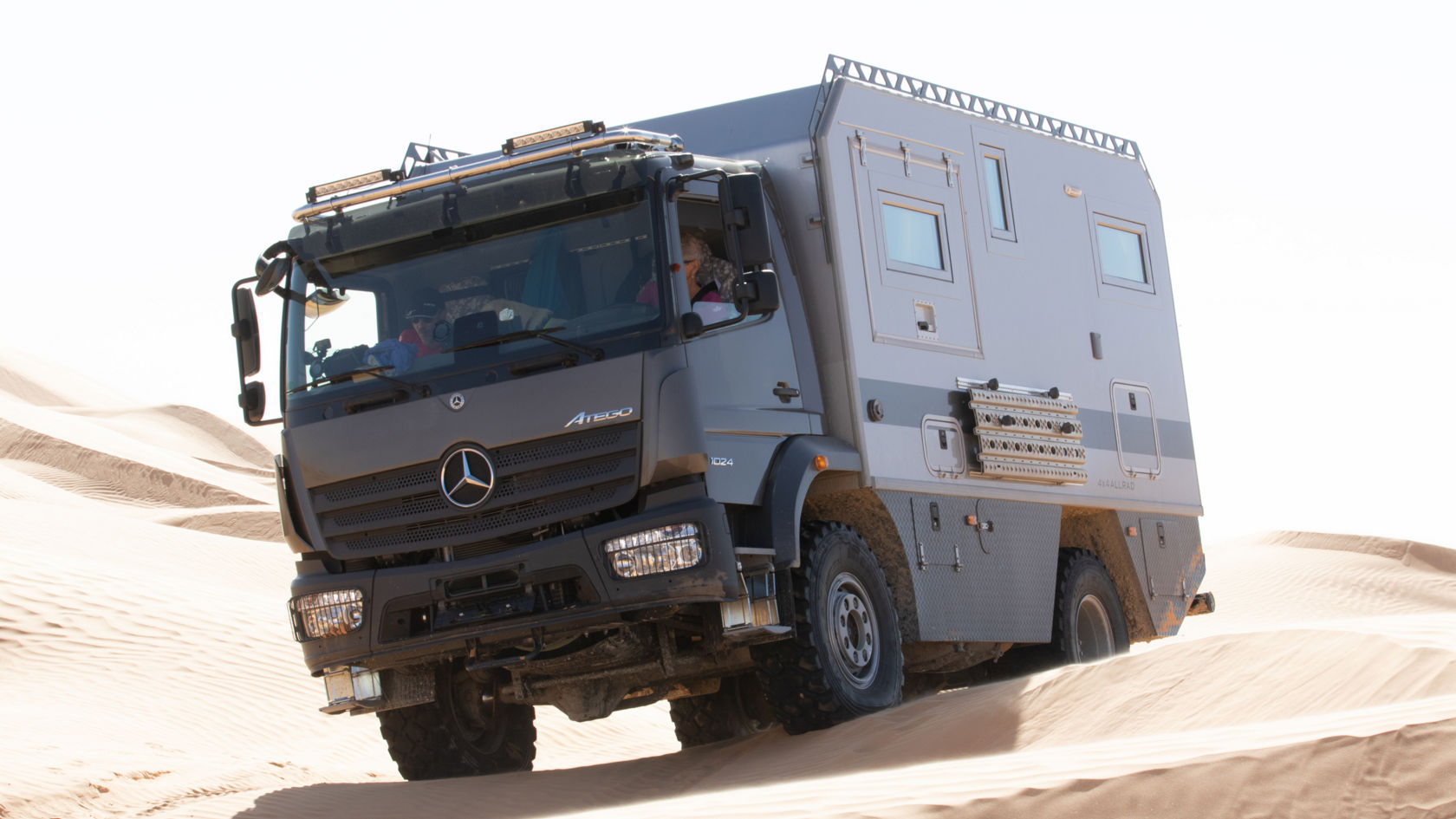 Das Bild zeigt einen grauen Atego mit einem Expeditionsmobil-Aufbau, der in einer Wüstenlandschaft unterwegs ist. Das Fahrzeug ist mit zusätzlichen Scheinwerfern auf dem Dach ausgestattet und scheint eine Sanddüne zu erklimmen. Der Himmel ist hell und sonnig.