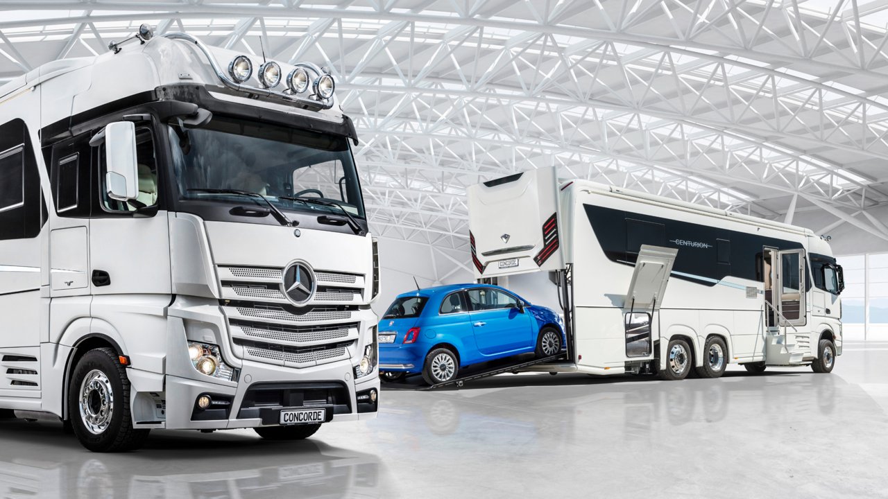 Das Bild zeigt zwei weiße Concorde Centurion Reisemobile in einer Halle mit einem modernen, gitterartigen Dach. Zwischen den beiden Reisemobilen befindet sich ein blauer Fiat 500, der in einer Art Garage im hinteren Teil des rechten Reisemobils geparkt ist. Die Reisemobile sind luxuriös ausgestattet und basieren auf einem Actros Chassis.