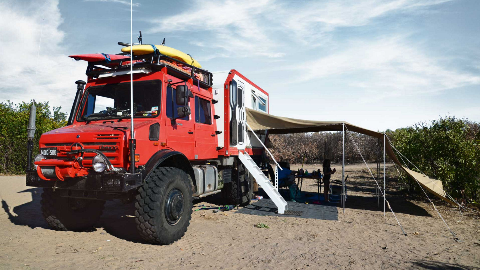 Das Bild zeigt einen roten Unimog mit einem Wohnmobilaufbau, der auf einem sandigen Untergrund steht. Auf dem Dach des Fahrzeugs sind Kajake und ein Fahrrad befestigt. Eine Markise ist ausgefahren und bietet Schatten für einen Campingbereich mit Tisch und Stühlen. Im Hintergrund sind Bäume und ein bewölkter Himmel zu sehen.