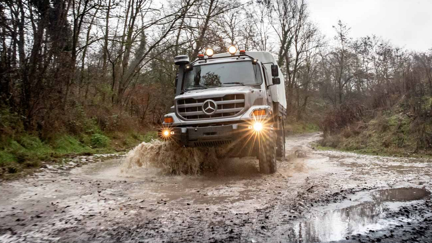 Nuotraukoje pavaizduotas pilkos spalvos „Zetros“ su kemperio kėbulu, važiuojantis per didelę balą ant klampaus kelio. Važiuodama per kliūtį, transporto priemonė taško vandenį. Fone matyti medžiai ir krūmai, supantys kelią. Dangus yra debesuotas ir atrodo, kad diena yra lietinga.