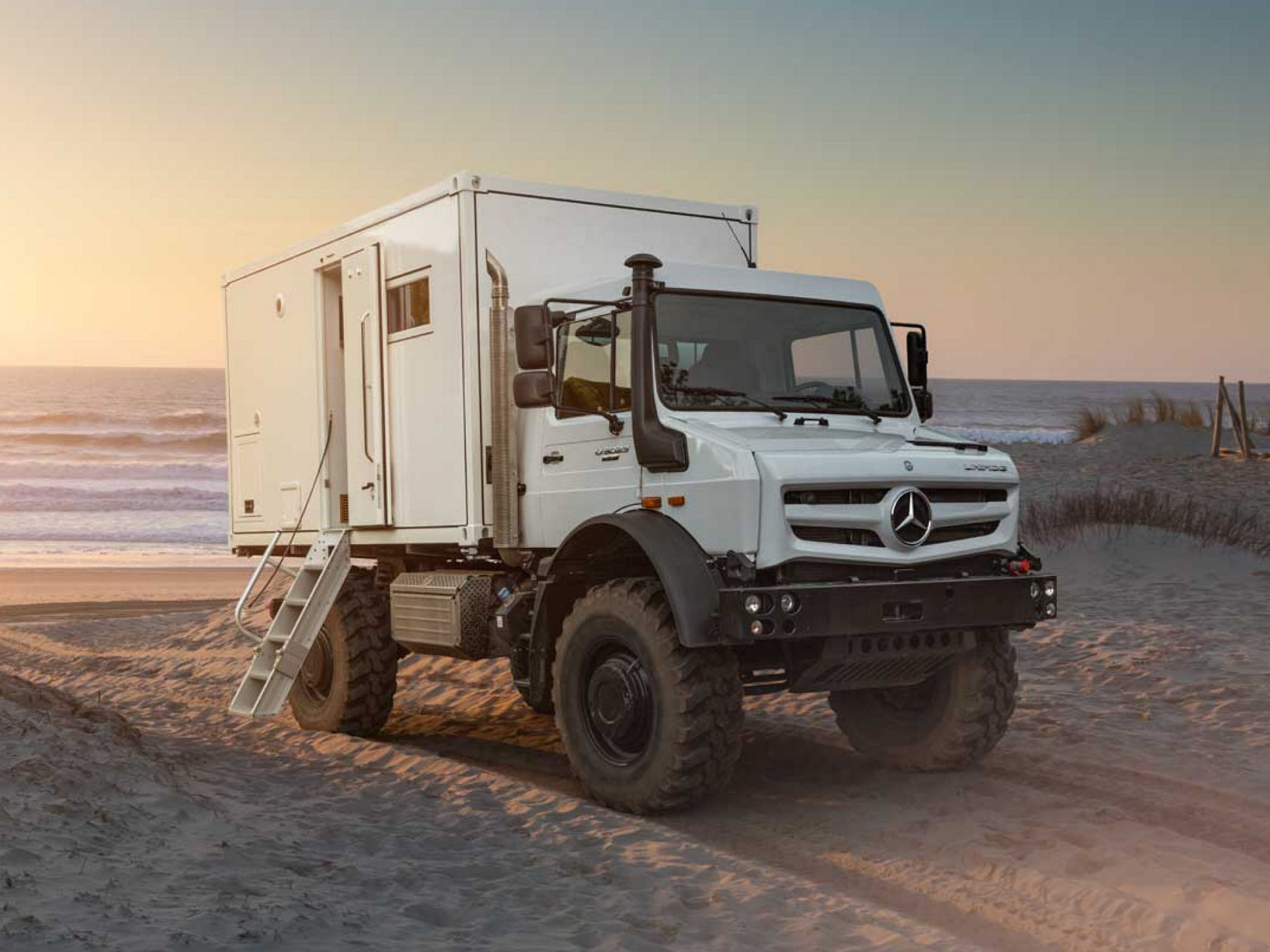 Das Bild zeigt zwei weiße Concorde Centurion Reisemobile in einer Halle mit einem modernen, gitterartigen Dach. Zwischen den beiden Reisemobilen befindet sich ein blauer Fiat 500, der in einer Art Garage im hinteren Teil des rechten Reisemobils geparkt ist. Die Reisemobile sind luxuriös ausgestattet und basieren auf einem Actros Chassis.