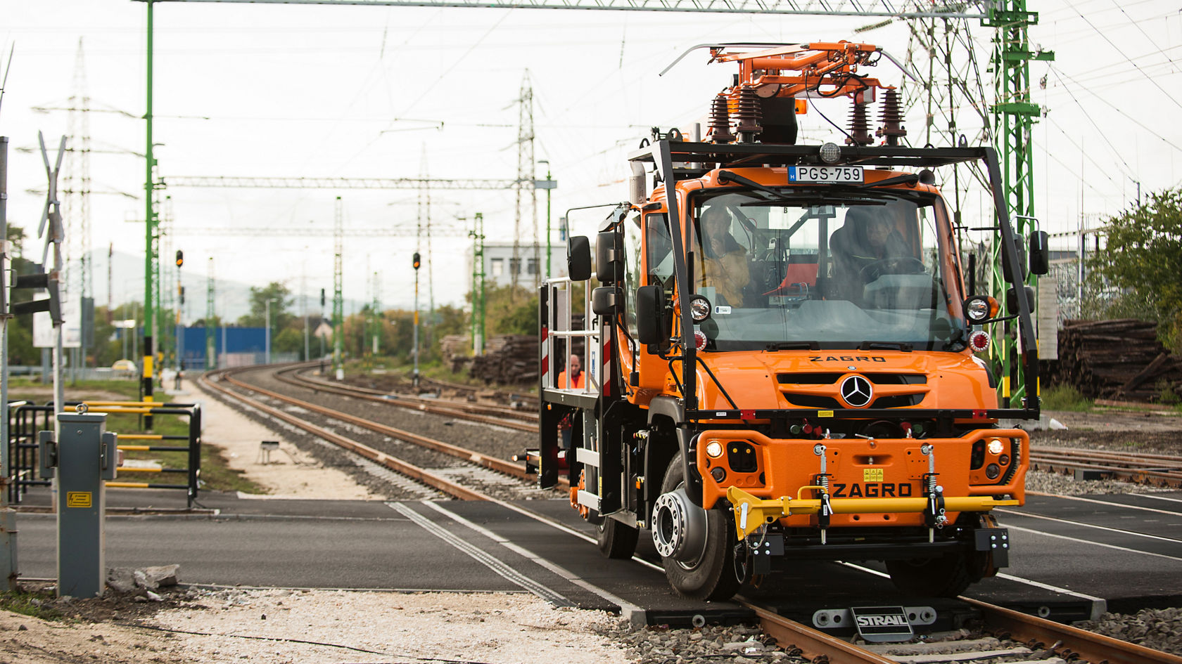 Oranži värvi Unimog õhuliinivarustusega, rööbastel raudtee piirkonnas, kus on signaalsüsteemid ja elektrimastid.