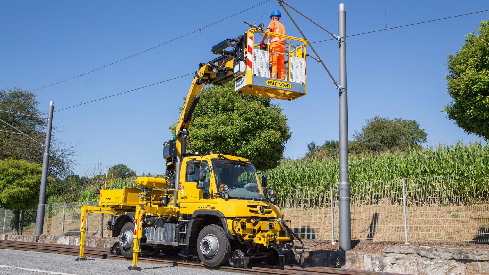 黄色 Unimog 带工作平台，用于农村环境中轨道上的架空线路维护作业。