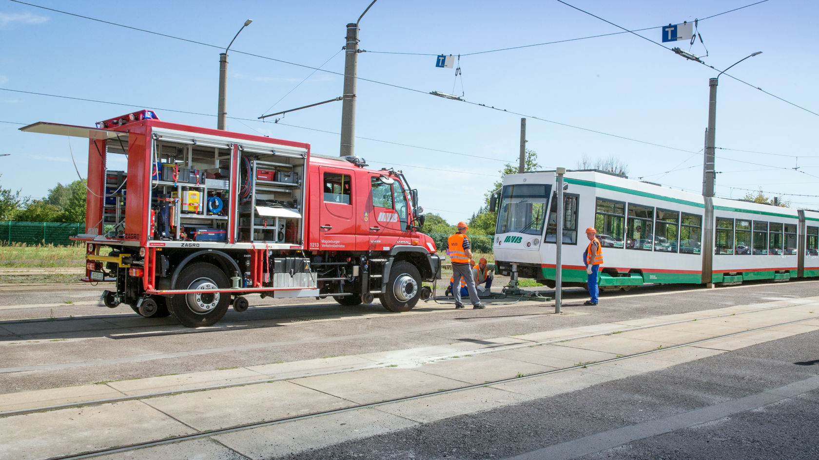 Punane Unimog seisab tänaval valge-rohelise trammi kõrval. Oranžides vestides töötajaid on näha sõidukite vahel, tõenäoliselt hooldustööde ajal. Veokil on avatud tagakülg seadmetega. Elektrijuhtmed ulatuvad üle sõidukite.