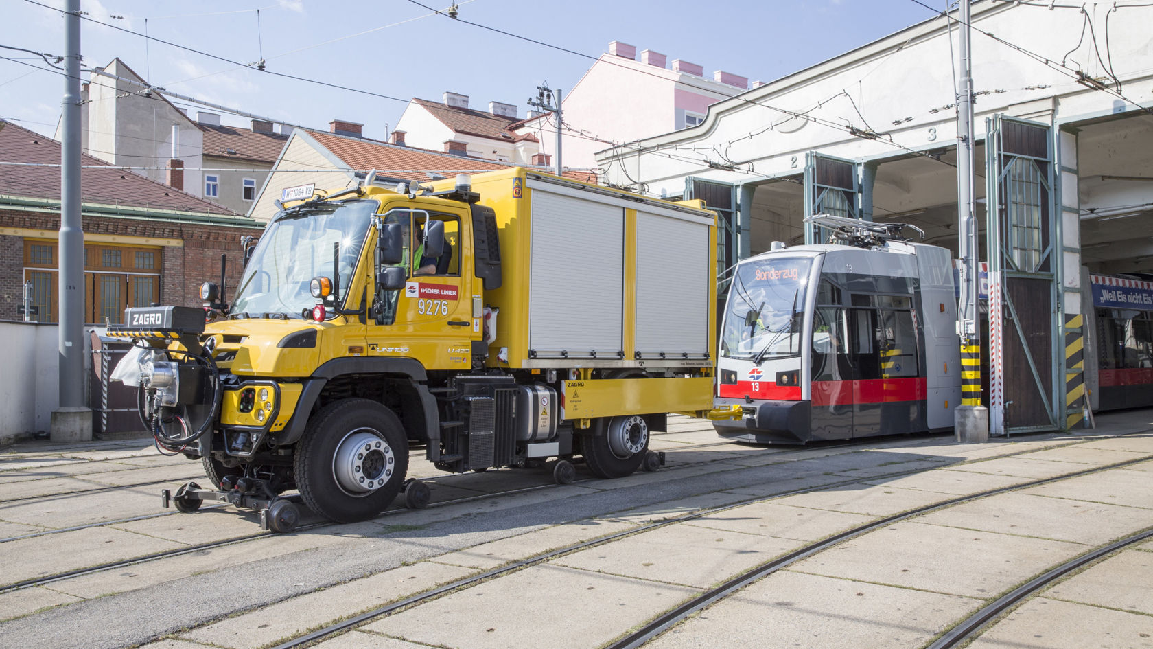 Kollane Unimog rööbastel, pargitud trammi ja depoohoone ette linnakeskkonnas.