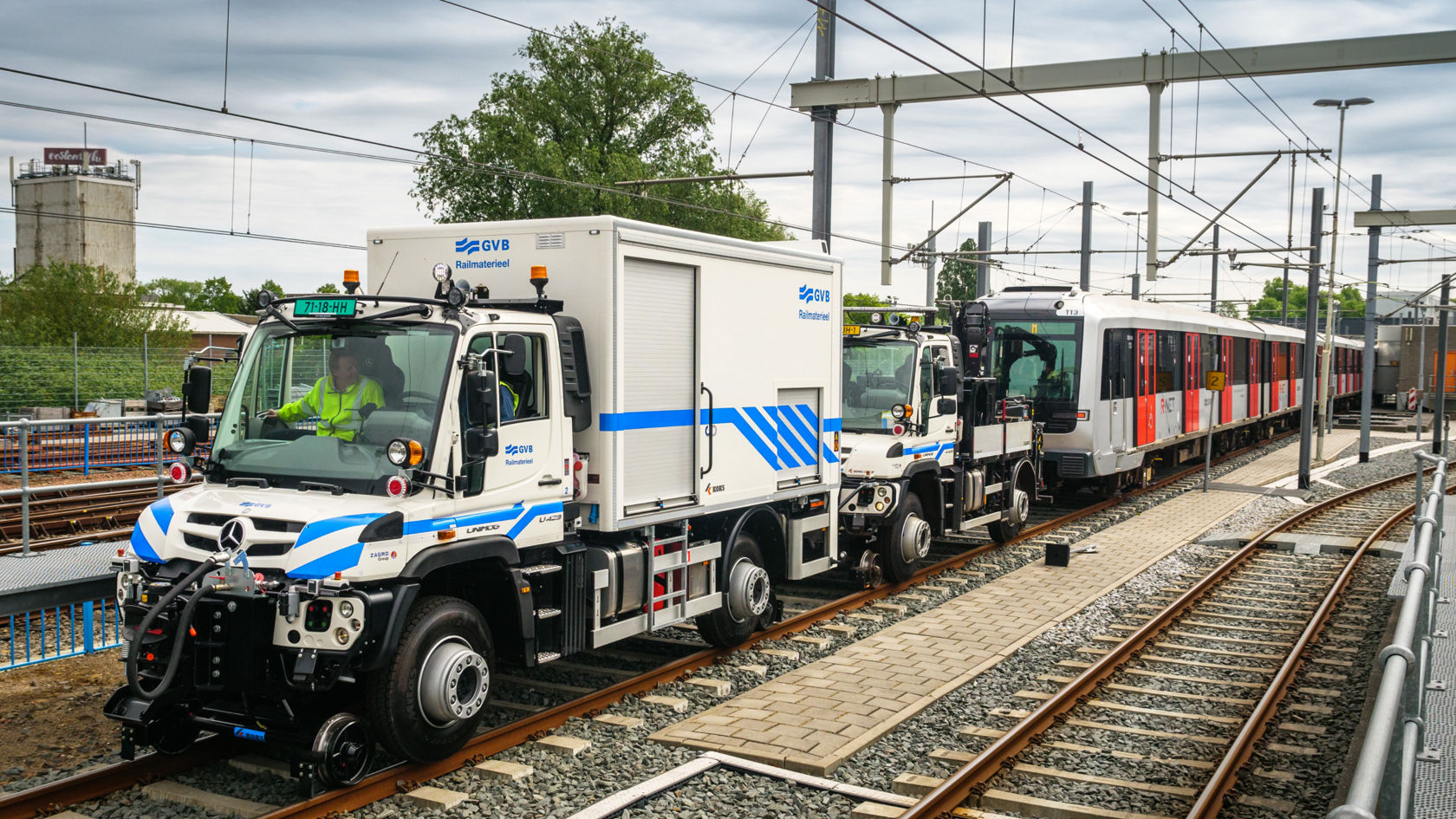 Kaks Unimogi kirjega rööbastel, punase ja valge trammi kõrval raudtee depoos.