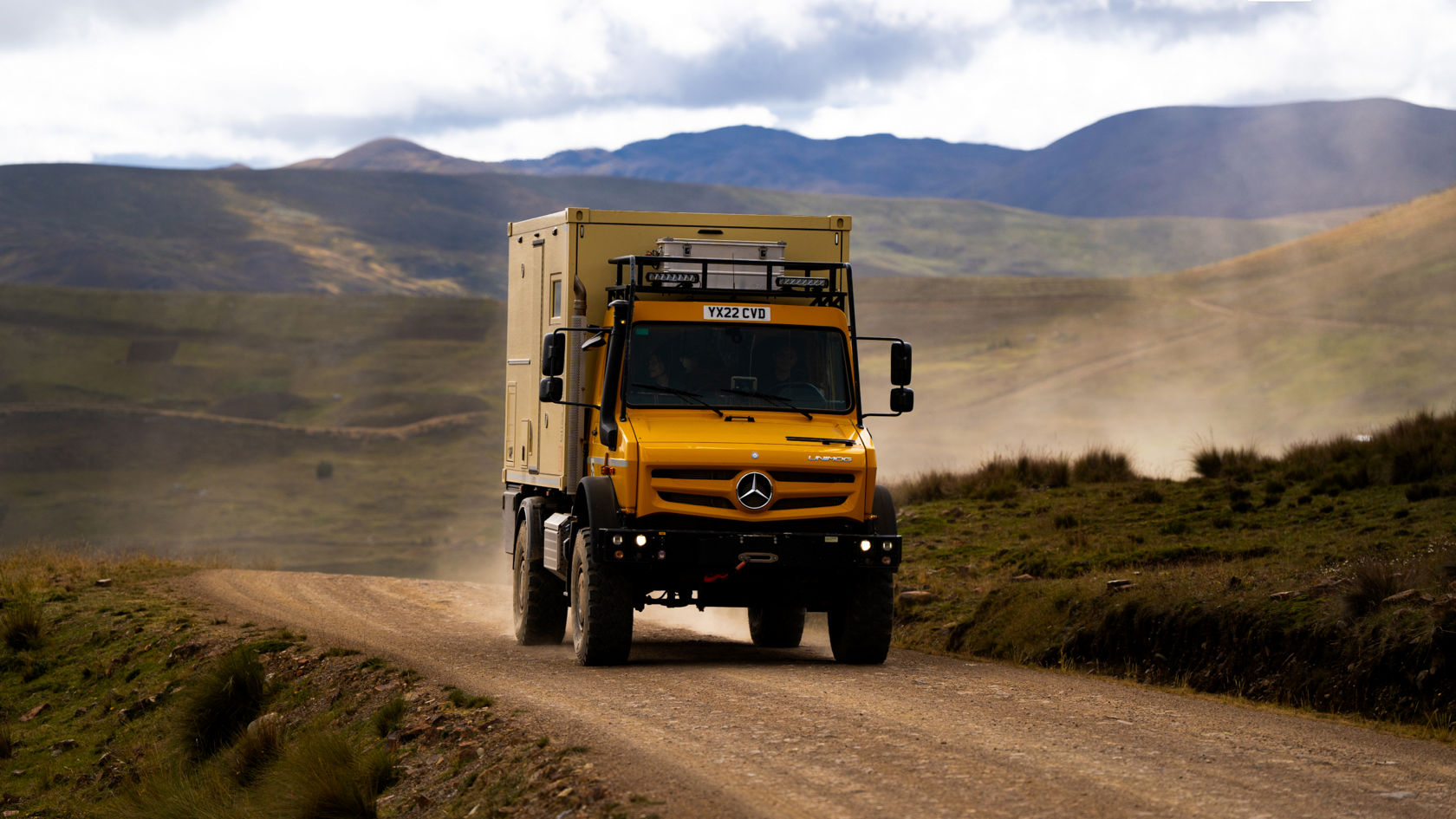 Das Bild zeigt einen gelben Unimog mit einem Aufbau, der wie ein Expeditionsmobil aussieht. Das Fahrzeug fährt auf einer unbefestigten Straße durch eine hügelige Landschaft. Im Hintergrund sind Berge und ein bewölkter Himmel zu sehen. Der Unimog ist mit einem Dachgepäckträger und zusätzlichen Scheinwerfern ausgestattet.