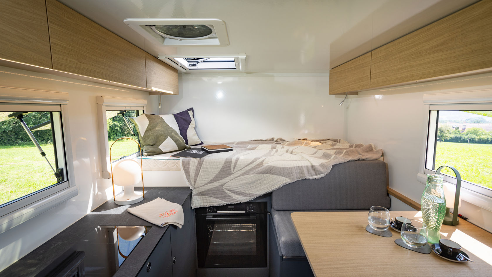 Das Bild zeigt das Innere eines modernen Wohnmobils oder Wohnwagens. Der Raum ist hell und sauber, mit weißen Wänden und hellen Holzmöbeln. Es gibt ein Bett mit einer grauen Tagesdecke und einem dekorativen Kissen, einen kleinen Esstisch mit Gläsern und Tassen sowie eine Küchenzeile mit einer schwarzen Arbeitsplatte. Ein Ofen ist unter dem Bett eingebaut. Fenster lassen Tageslicht herein und bieten einen Blick auf eine grüne Landschaft.
