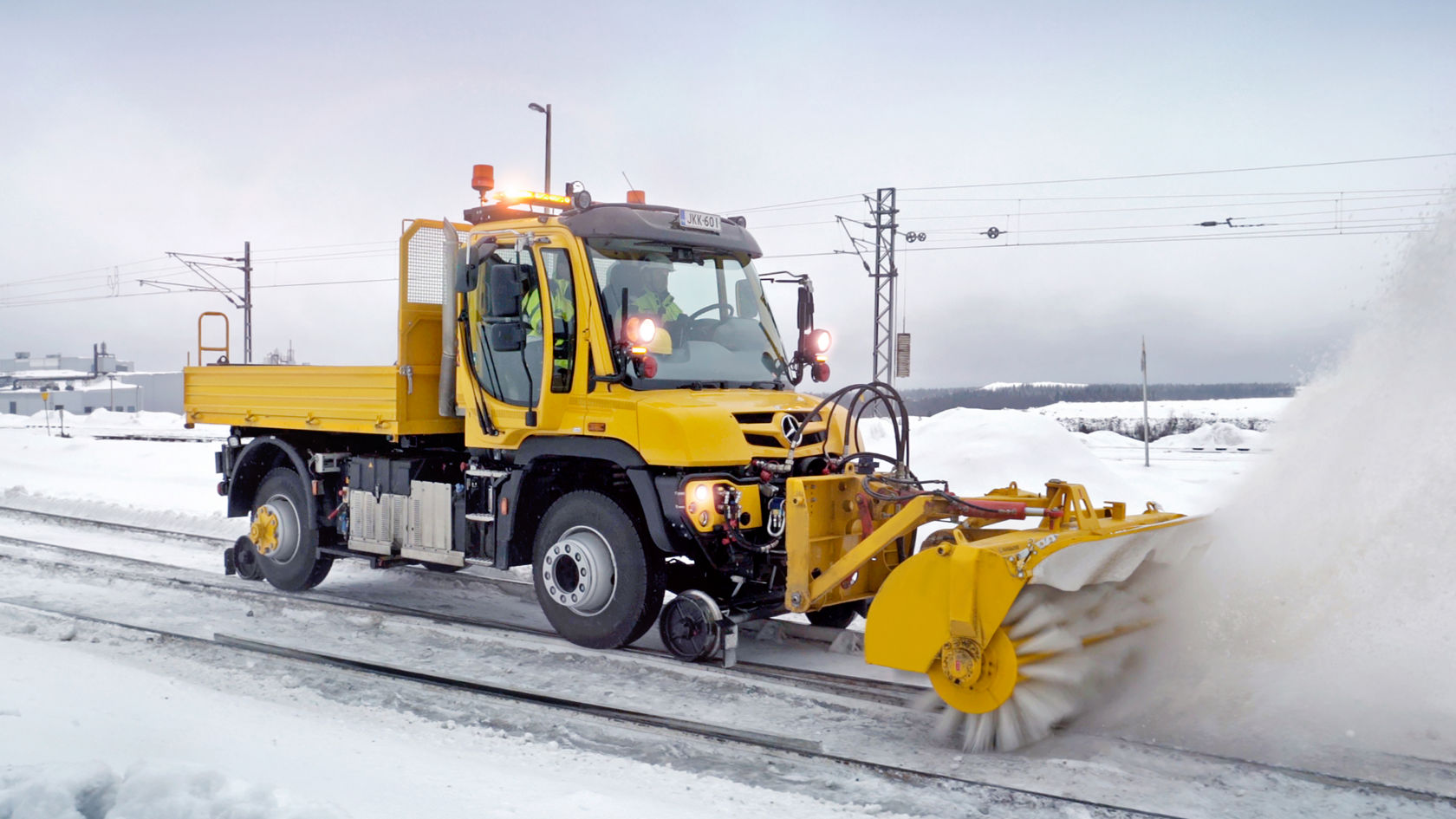 Lumepuhastusseadmega kollane Unimog talvises keskkonnas lumega kaetud rööbastel.