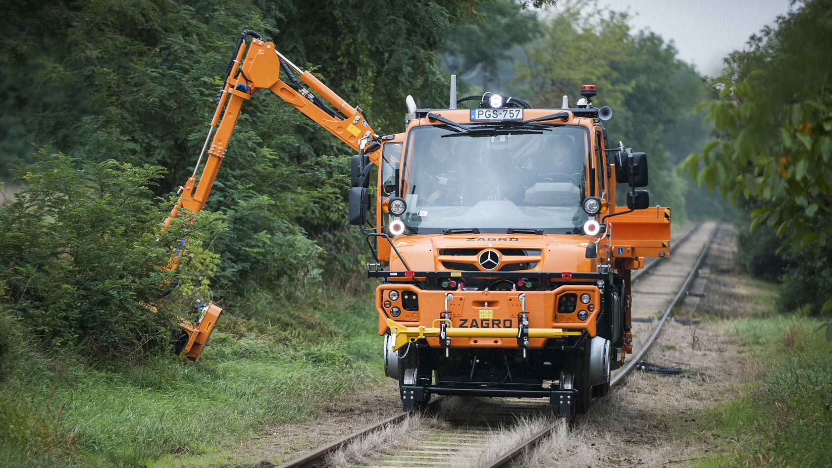 Spetsiaalse varustusega oranž Unimog sõidab rööbastel ja lõikab külgmise noolega piki teed põõsaid.
