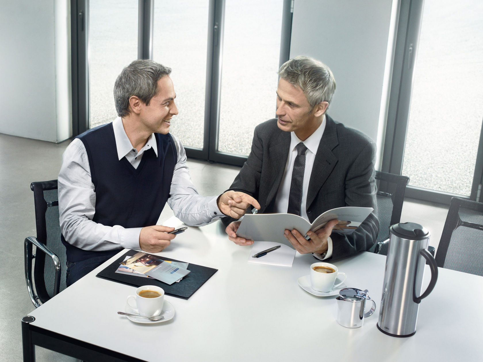 Twee zakenmensen zitten aan een tafel, bespreken documenten en drinken koffie.