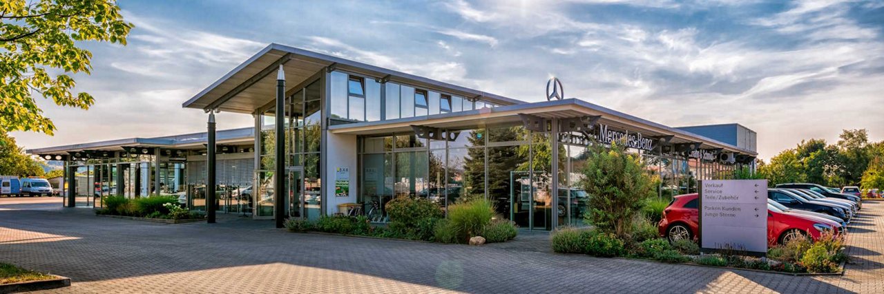 Außenansicht des Mercedes-Benz Autohauses Hahn & Schmidt in Radebeul bei sonnigem Wetter. Das moderne Gebäude mit großer Glasfront ist von Grünflächen umgeben, vor dem Eingang stehen mehrere geparkte Fahrzeuge.