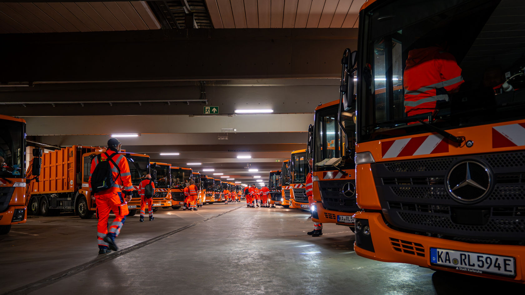 Eine Flotte von Econic Entsorgungsfahrzeugen steht in einer großen Halle eines städtischen Fuhparks. Dazwischen stehen Müllwerker an den Fahrzeugen.