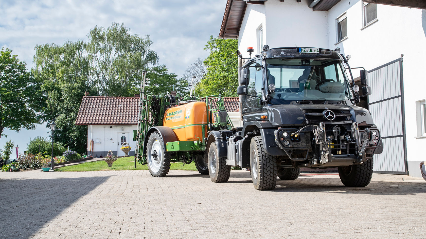 Un Unimog Mercedes‑Benz est stationné sur une cour pavée, tandis qu'il tracte une remorque de pulvérisation aux couleurs vives. En arrière-plan, on aperçoit des arbres et un bâtiment.