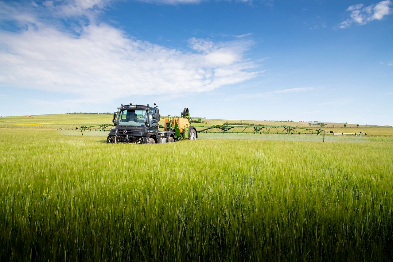 Un tracteur Mercedes‑Benz pulvérise un champ verdoyant, entouré de collines et d'un ciel bleu parsemé de quelques nuages. L'entretien des machines agricoles est visible.