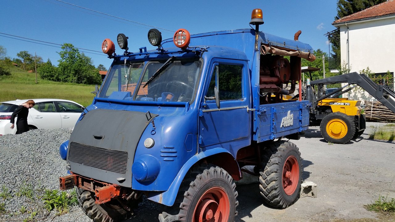 Blauer Mercedes-Benz Unimog mit mehreren Scheinwerfern, geparkt auf einem Gelände mit Kies und einer gelben Maschine im Hintergrund.