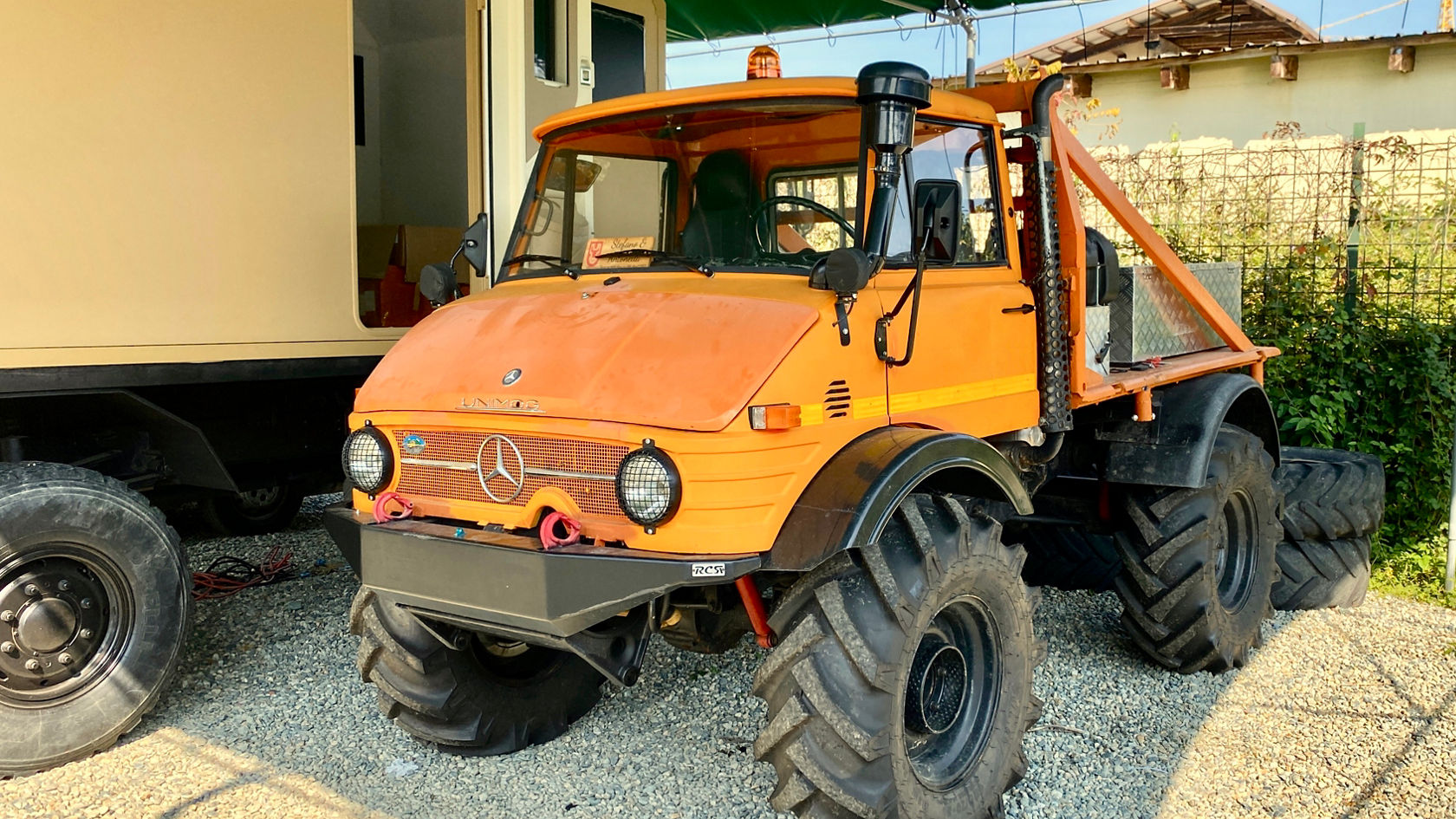 Ein orangefarbener Mercedes-Benz Unimog steht auf einem Kiesplatz, mit großen, robusten Reifen und einer offenen Ladefläche. Der Fahrersitz und das Armaturenbrett sind gut sichtbar.