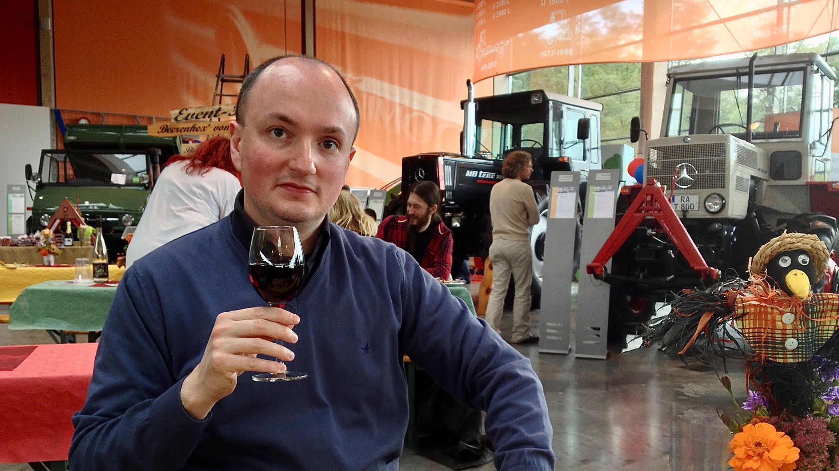 Ein Mann hält ein Glas Rotwein in der Hand, während er in einem Raum mit mehreren Traktoren und einem Unimog von Mercedes-Benz im Hintergrund sitzt.
