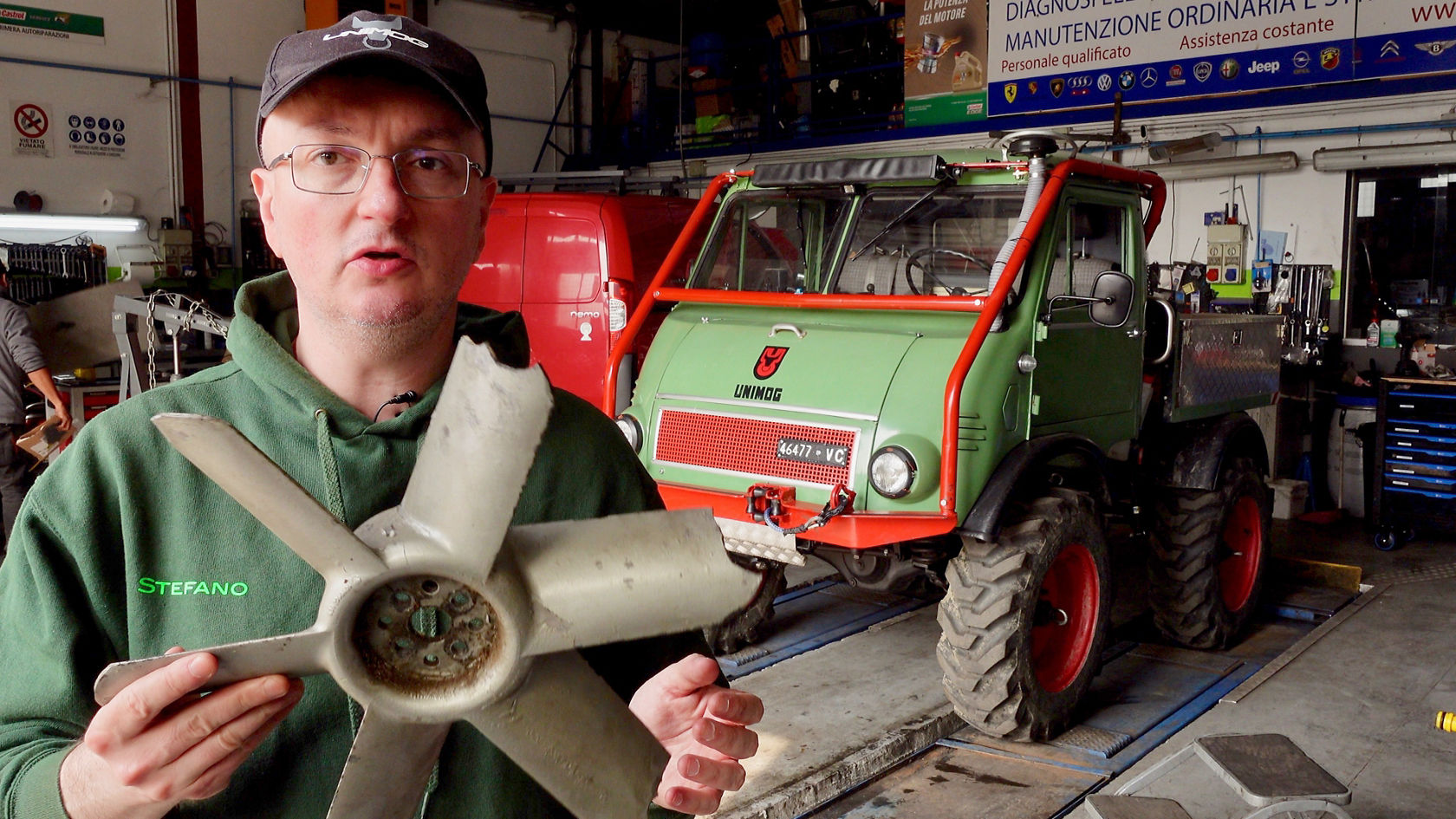 Ein Mann hält einen Kühlerlüfter in der Hand, während ein grüner und roter Unimog von Mercedes-Benz im Hintergrund sichtbar ist, in einer Werkstatt.