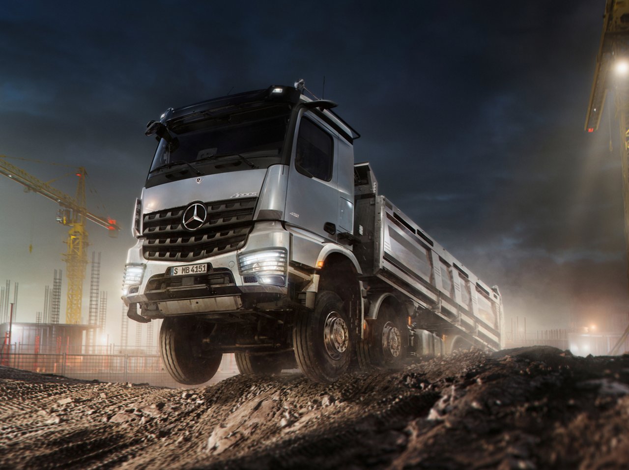 Ein silberner Mercedes-Benz Baustellen-LKW fährt bei Nacht über unebenes Gelände auf einer Baustelle. Im Hintergrund sind Baukräne zu sehen.