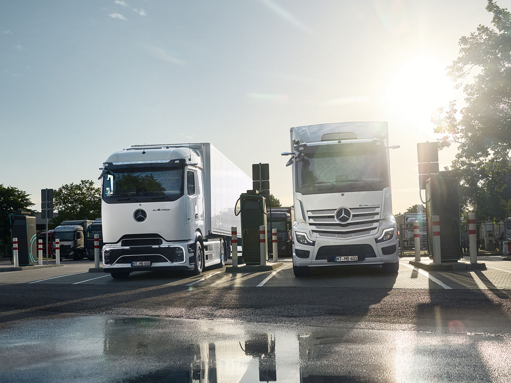 Zwei weiße eTrucks von Mercedes-Bent Trucks stehen nebeneinander an Ladestationen auf einem nassen Parkplatz, der die Fahrzeuge und die Umgebung spiegelt. Die Sonne scheint hell am Himmel und erzeugt eine starke Blendung und lange Schatten, während Bäume und Gebäude im Hintergrund zu sehen sind.
