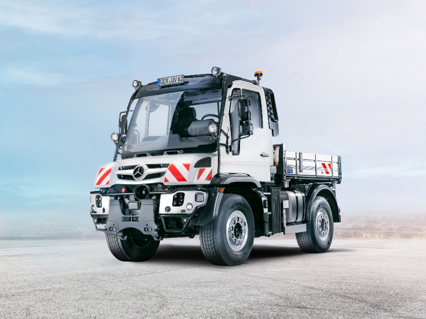 Ein weißer Unimog steht auf einer leeren Straße. Der Lkw hat eine offene Ladefläche und ein großes Fahrerhaus.