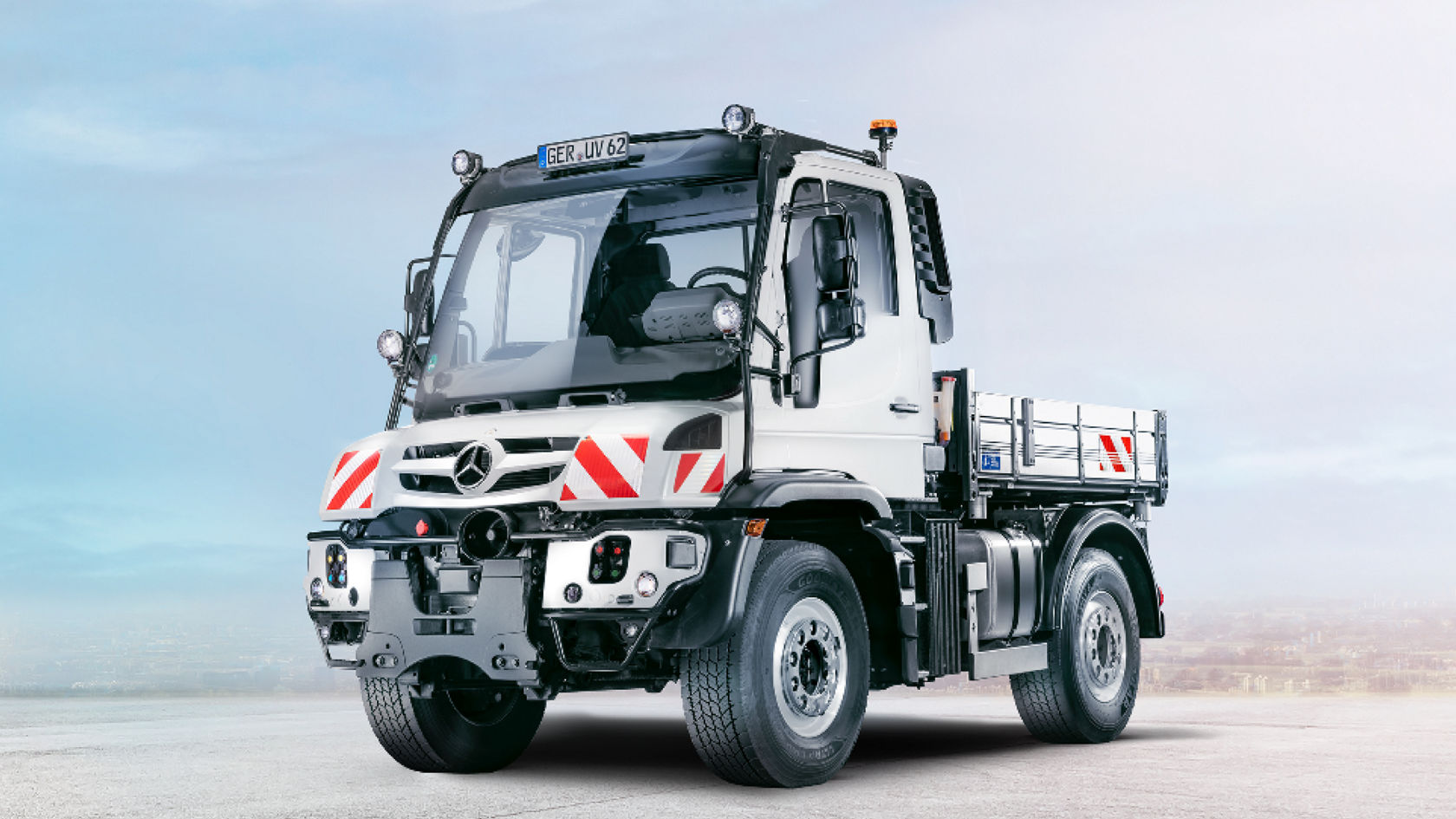 Ein weißer Unimog steht auf einer leeren Straße. Der Lkw hat eine offene Ladefläche und ein großes Fahrerhaus.