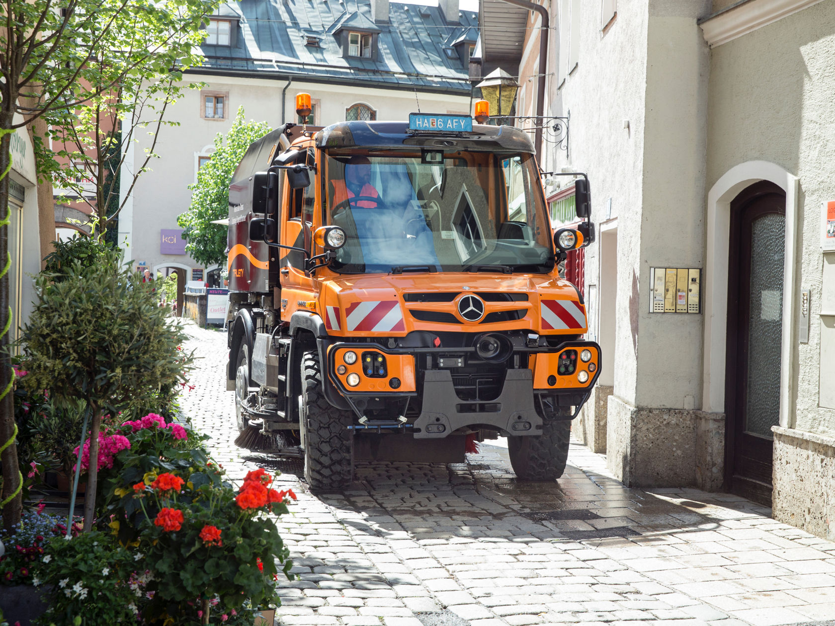 Ein oranger Unimog Geräteträger im Kommunal-Einsatz fährt durch eine schmale Gasse mit Kopfsteinpflaster. Links sind Blumenbeete zu sehen.