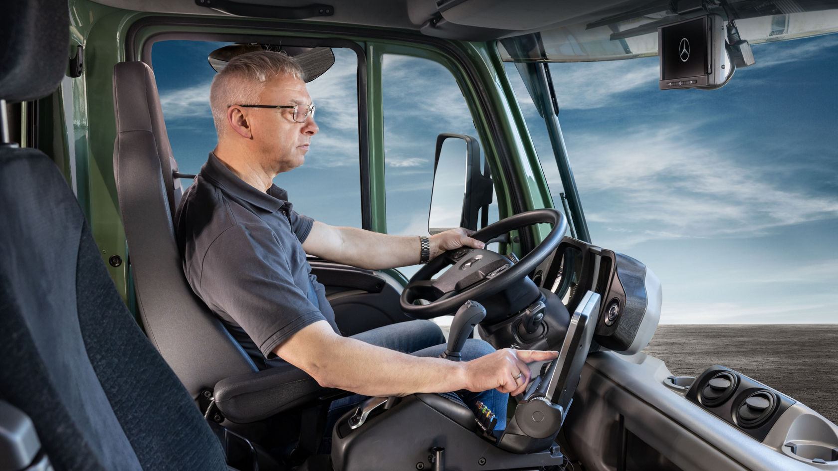 Un homme est assis au volant d'un Unimog et utilise un panneau de contacteurs. Il porte une chemise bleue et des lunettes.