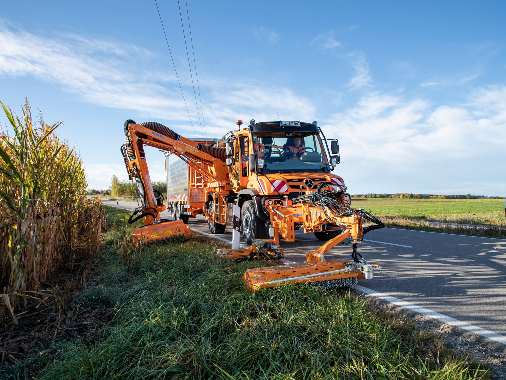 Un porte-outils Unimog orange avec faucheuse tond l'herbe sur le bord de la route. Le véhicule roule sur une route qui longe un champ.