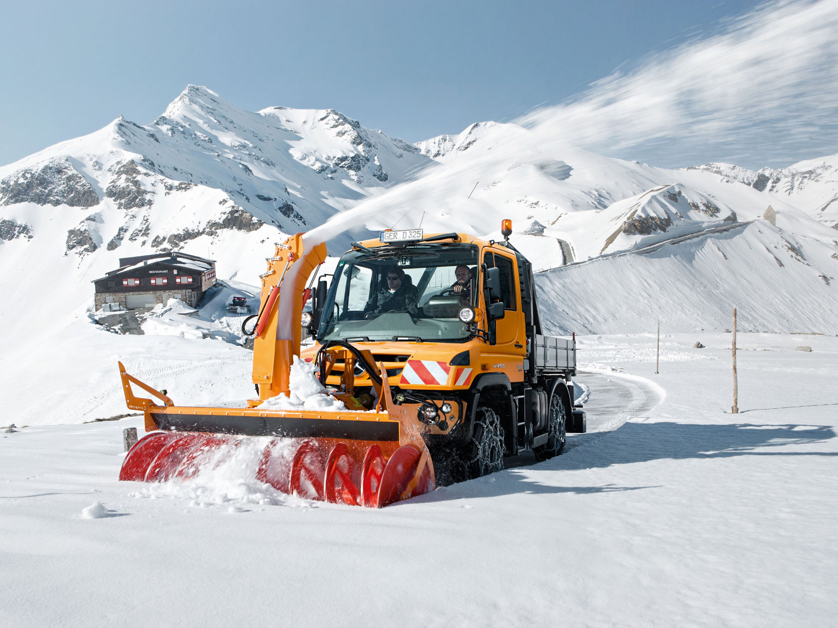 Un porte-outils Unimog orange déneige un chemin dans une région montagneuse enneigée.