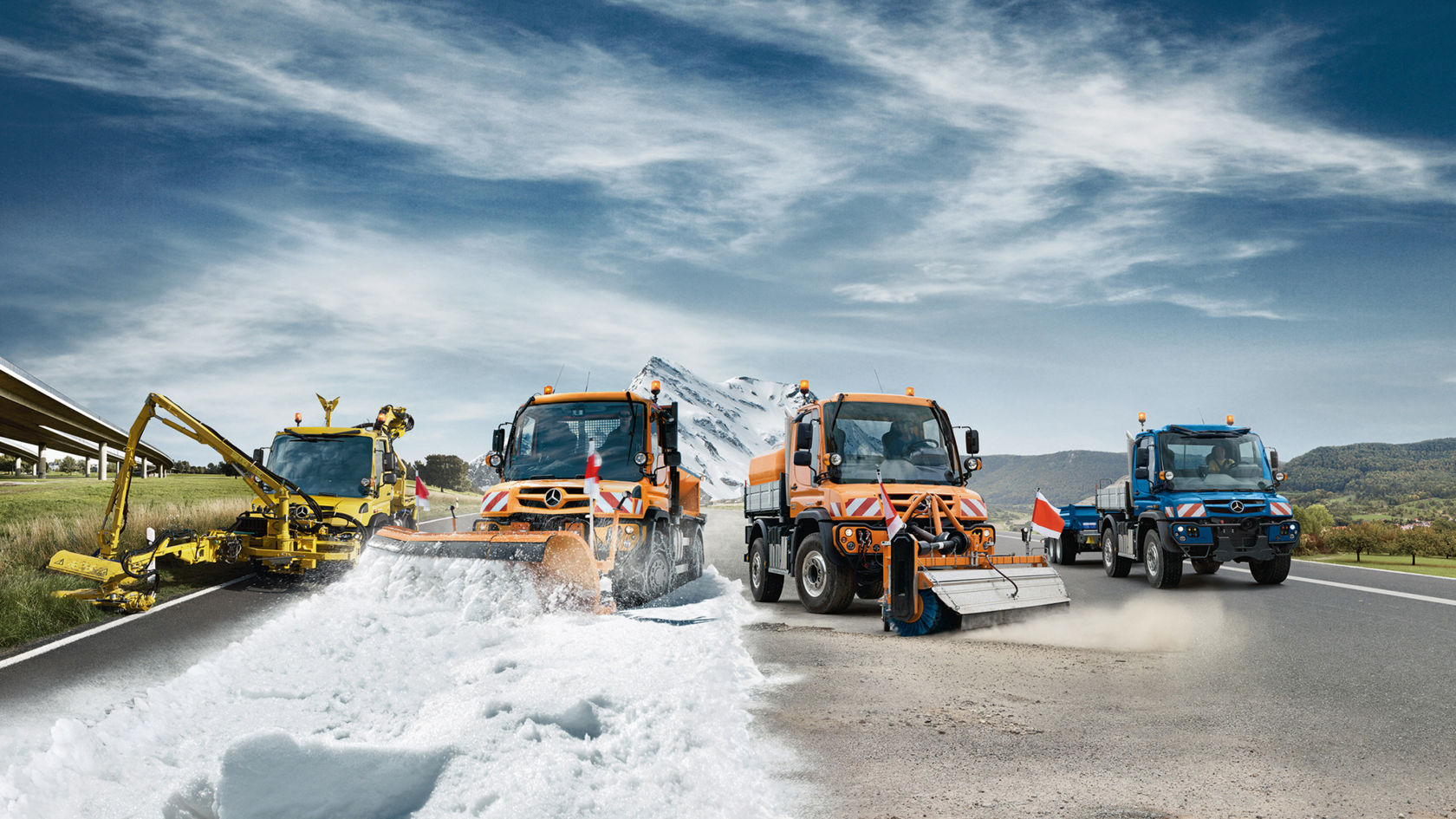 Vier Unimog werktuigdragers in verschillende kleuren en toepassingen staan naast elkaar op een brede geasfalteerde weg: winterdienst, straatreiniging, landbouw en transport. Op de achtergrond zijn groene weiden en bergen, een met sneeuw bedekte berg en een brug oer een snelweg te zien. 