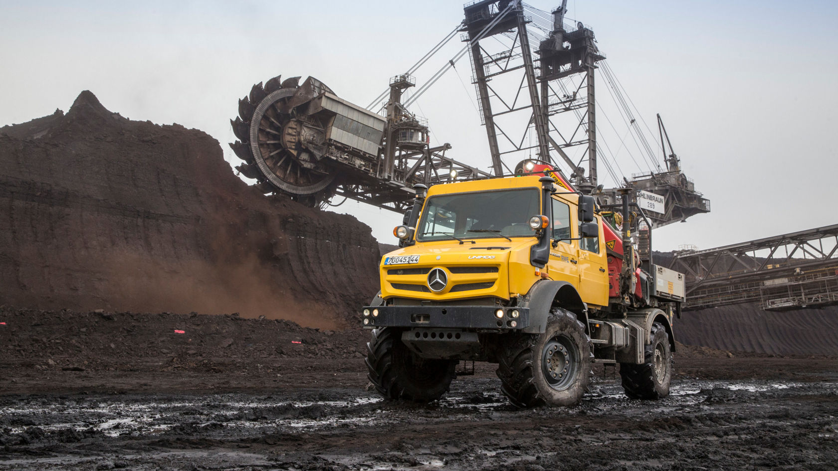 A yellow Mercedes-Benz commercial vehicle is standing in a coal pit, while a large mining excavator in the background is extracting coal. Dust and earth material are visible.