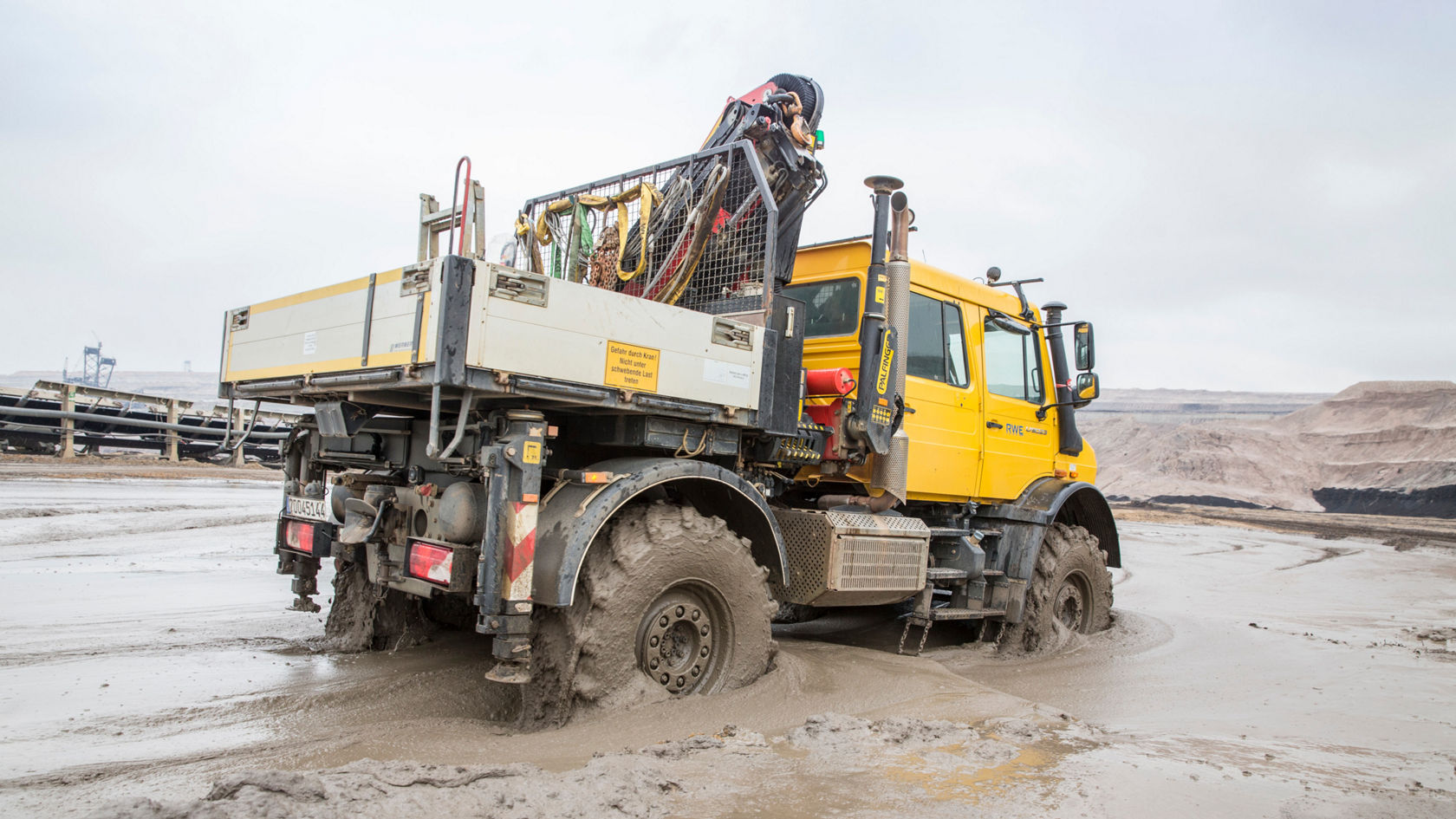 A Mercedes-Benz truck stands in muddy terrain. The cargo area is loaded with equipment. Typical open-cast mining work environments can be seen in the background.