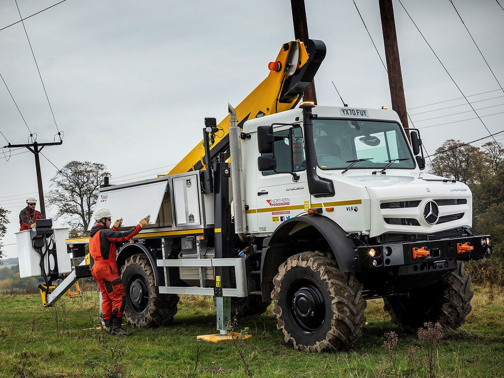 Ein Unimog mit einem Kranarm steht auf einem Feld. Ein Arbeiter befindet sich in einer Hebebühne, die an einem Strommast befestigt ist.