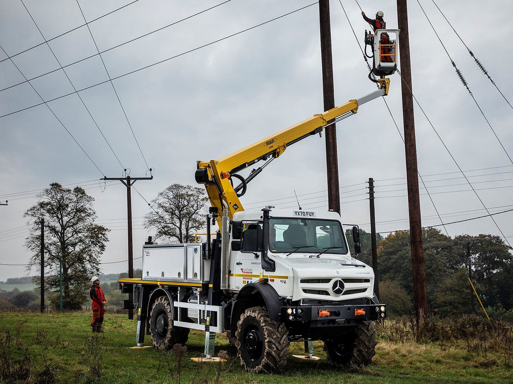 Ein Arbeitsfahrzeug, ein Mercedes-Benz Unimog, steht auf einer Wiese. Zwei Arbeiter in oranger Sicherheitskleidung sind dabei, eine Hebebühne vorzubereiten, um an eine Stromleitung zu gelangen.