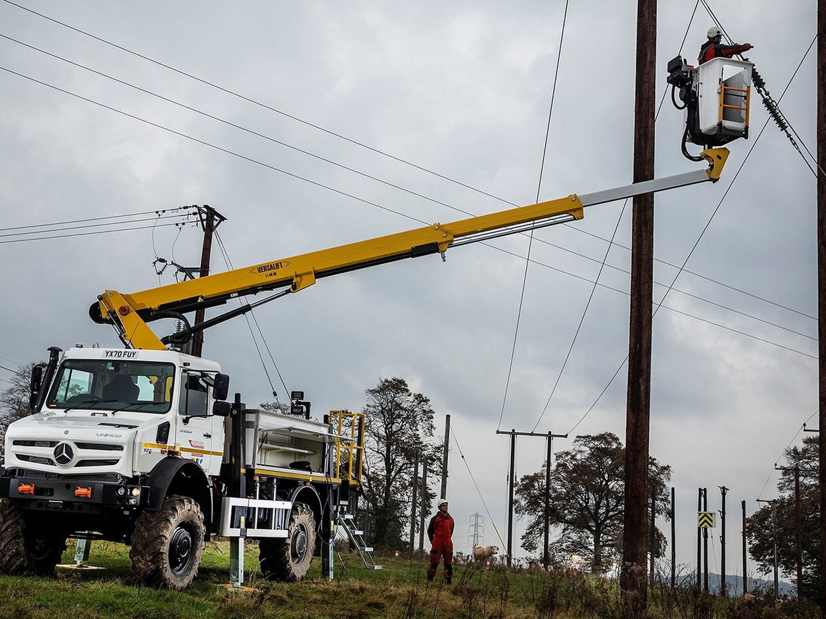 Ein Unimog mit Hubarbeitsbühne steht auf einem Feld. Ein Arbeiter in einem Sicherheitsgeschirr befindet sich in der Höhe an einem Strommast und führt Arbeiten durch.