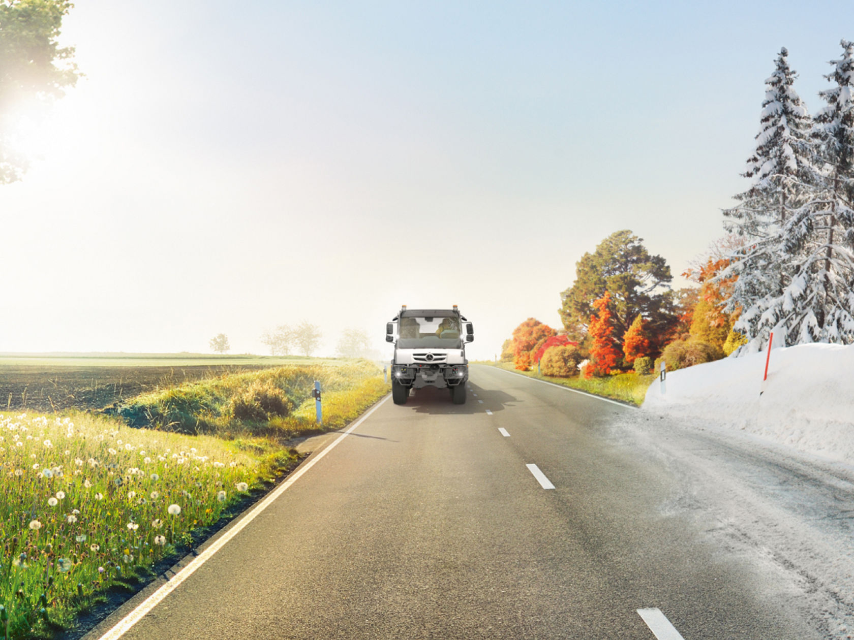 Ein Unimog fährt auf einer Landstraße, die durch eine geteilte Landschaft führt. Auf der linken Seite blüht eine frühlingshafte Wiese, während auf der rechten Seite winterliche Verhältnisse mit Schnee und verschneiten Bäumen herrschen.