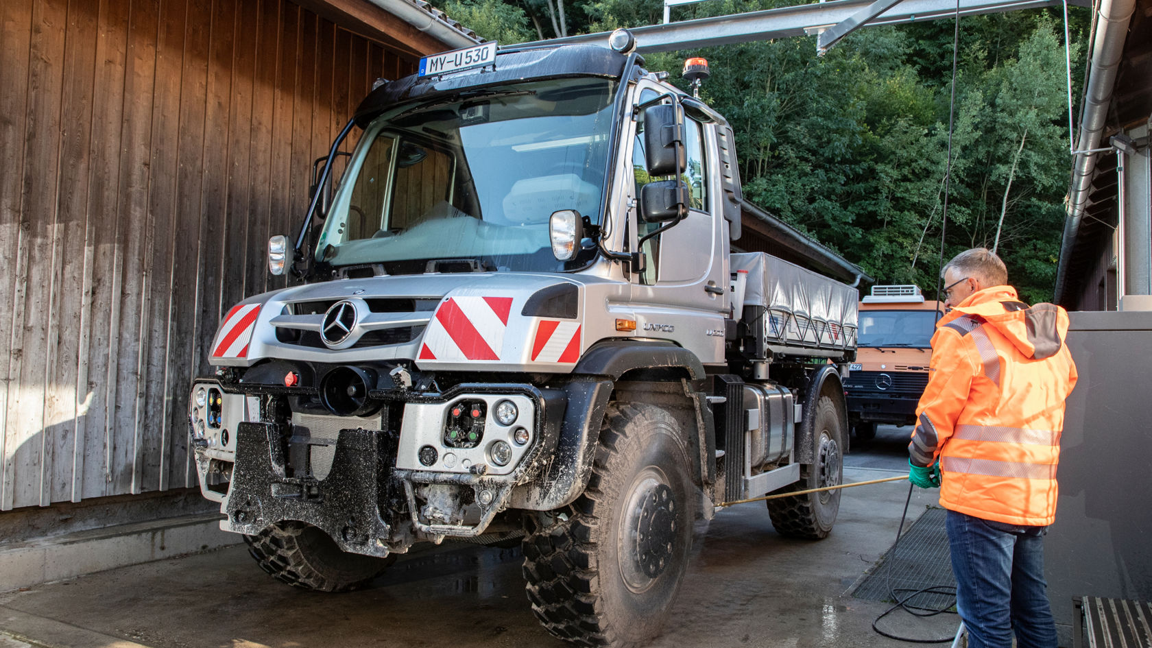 A man in an orange work jacket cleans a silver Unimog with a high-pressure cleaner. The truck is standing in front of a wooden building. In the background is another orange Unimog and trees.