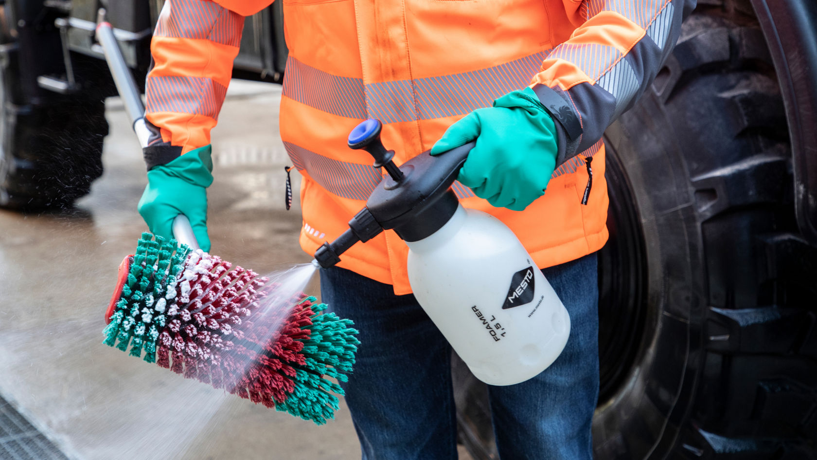 Een persoon in een oranje werkjas spuit reinigingsmiddel op een groen-rode borstel die aan een telescoopsteel is bevestigd.