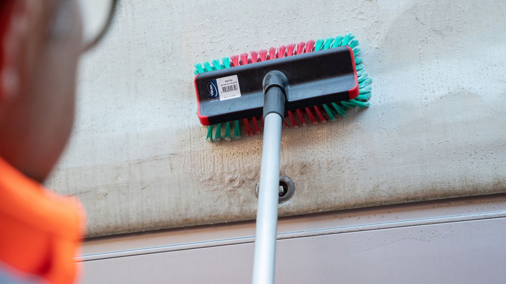 Une personne en veste de travail orange frotte la surface extérieure d'une bâche de camion avec une brosse montée sur un manche télescopique.