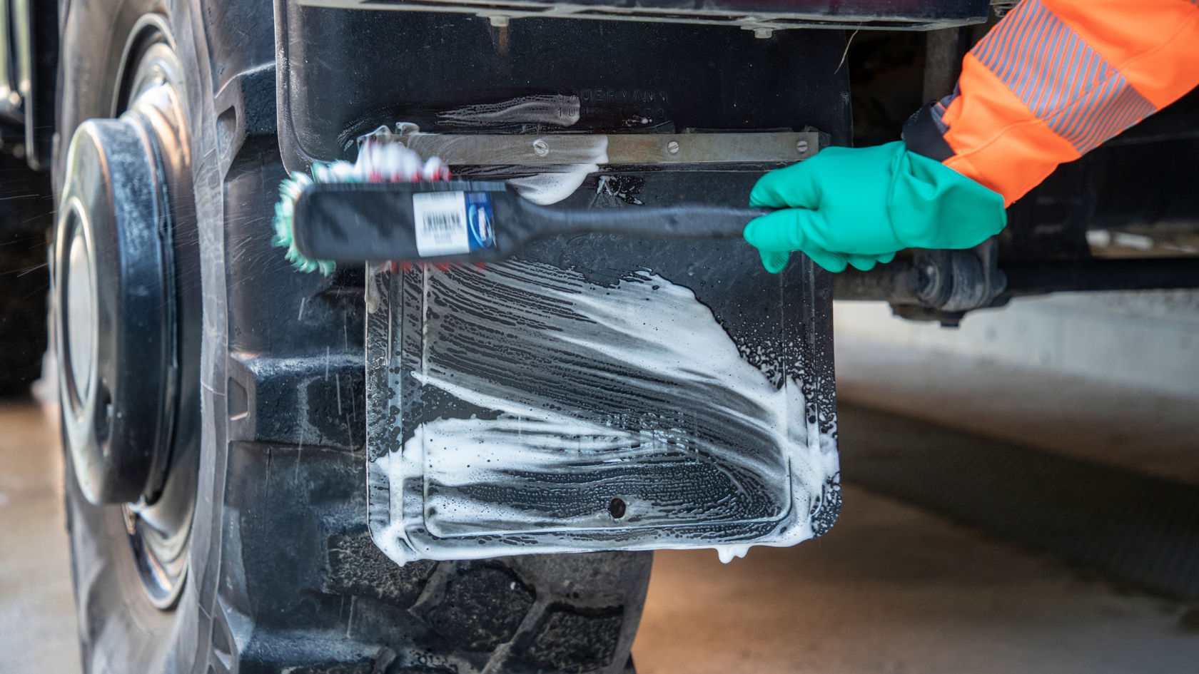 Une main en gant de caoutchouc vert frotte avec une brosse à main une tôle de protection noire sur le pneu d'un camion.