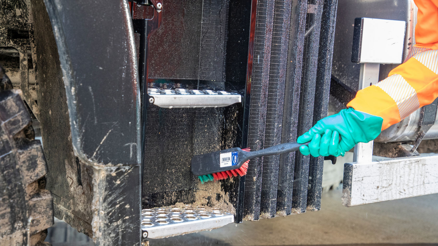 A hand wearing a green rubber glove cleans the steps of a Unimog using a hand brush. A few splashes of mud can be seen.