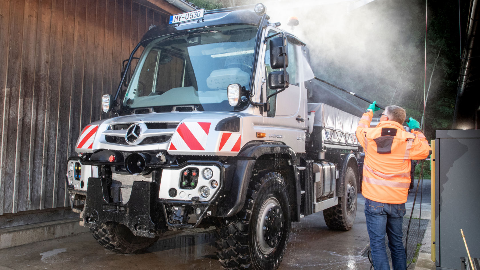 A man in an orange work jacket cleans a silver Unimog with a high-pressure cleaner. He is spraying the driver’s window. Spray can be seen above the truck.