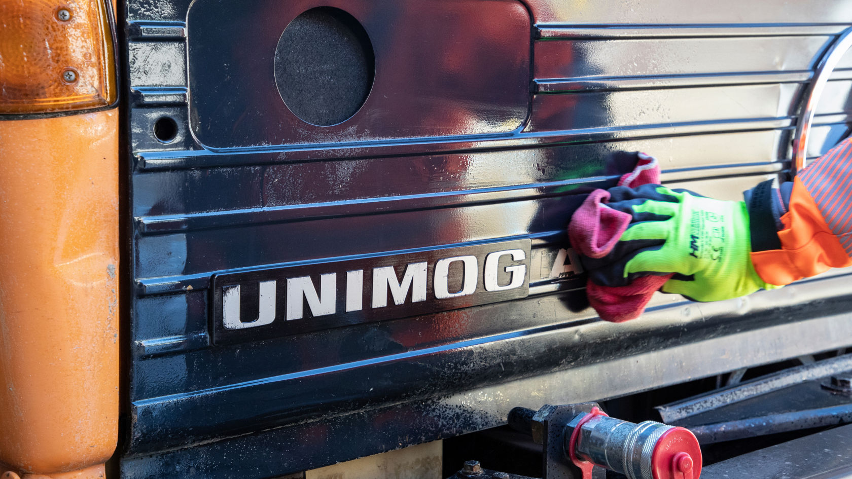 Eine Hand mit Arbeitshandschuh reinigt oder poliert den Unimog-Schriftzug an einem Fahrzeug.