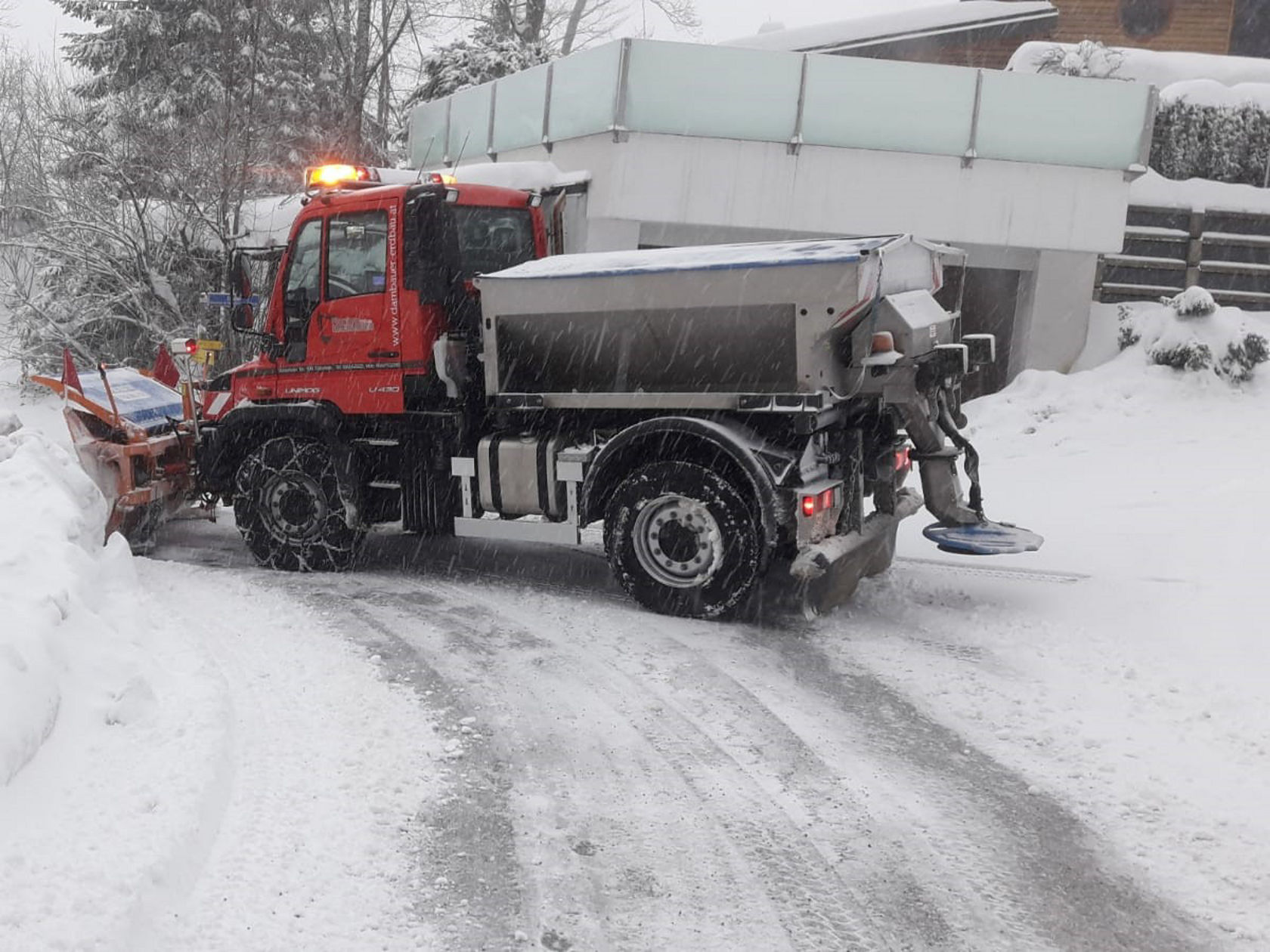 Schneeräumfahrzeug von Mercedes-Benz beim Räumen einer verschneiten Straße, während Schnee um das Fahrzeug herum angesammelt wird. Rote Farbe, mit Schneeschaufel und Streuer ausgestattet.