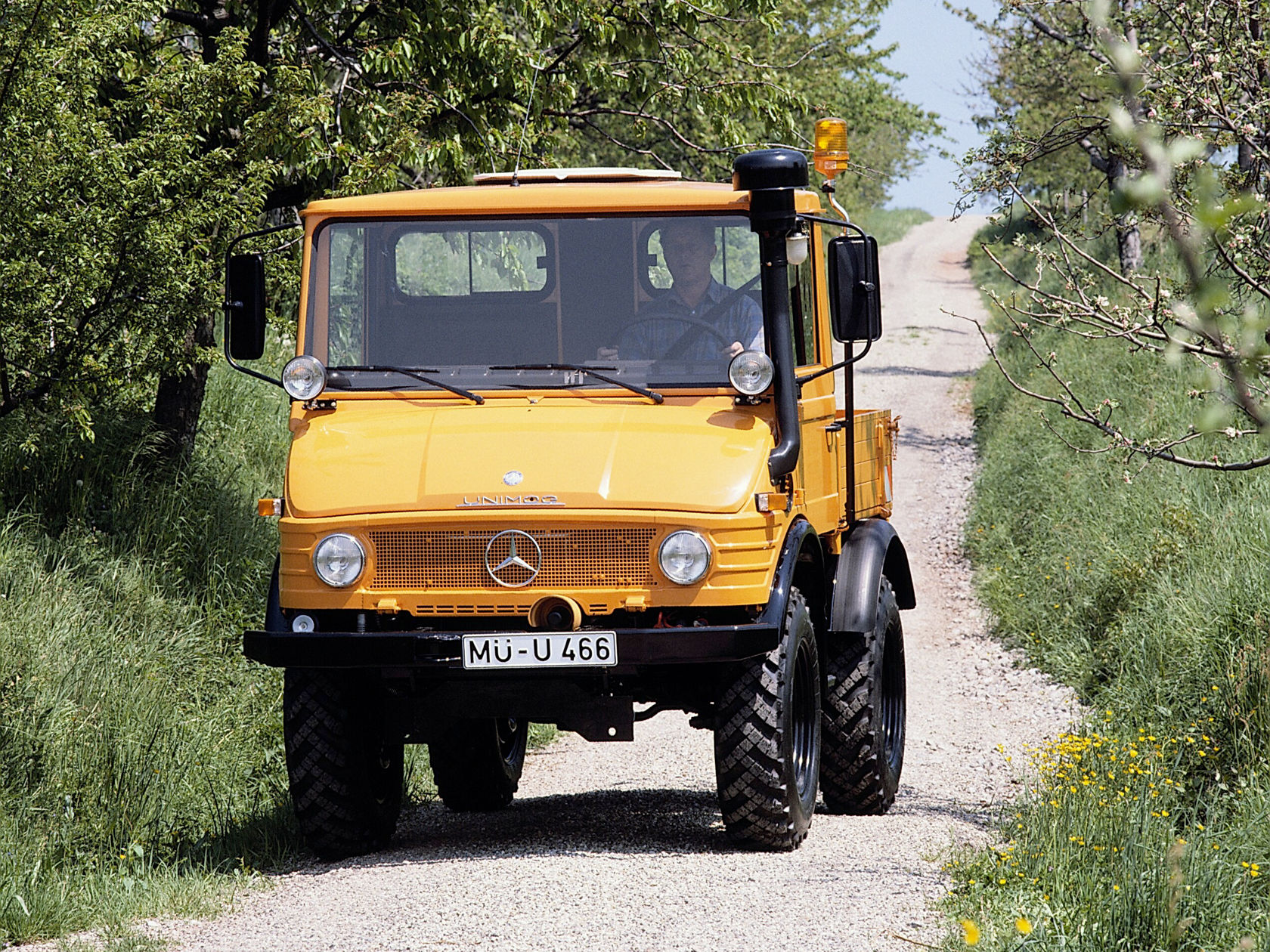 Een gele Mercedes-Benz Unimog rijdt op een smalle weg, omgeven door groene weiden en bomen, ideaal voor landbouw- en terreintoepassingen.