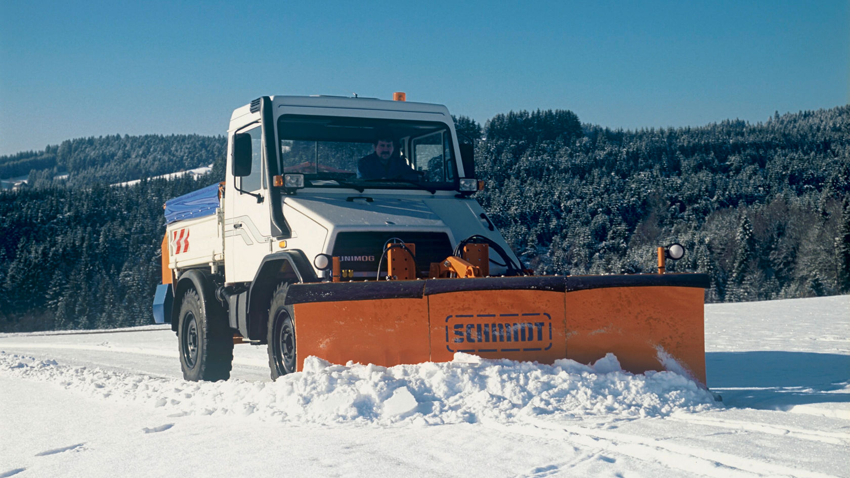 Een Mercedes-Benz Unimog met sneeuwploeg ruimt sneeuw van een besneeuwde weg in een winterlandschap met dennenbomen op de achtergrond.